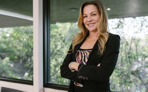 A smiling executive stands with arms crossed in front of a window overlooking trees
