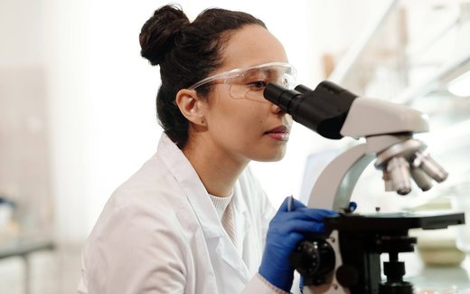 A scientist in a white lab coat, blue gloves and safety glasses looks through a microscope in a laboratory