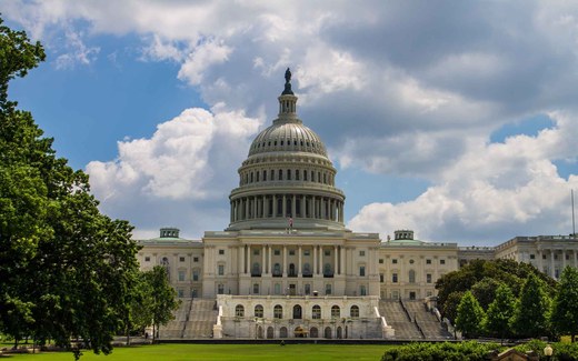 The dome of the U.S. Capitol rises against a partly cloudy blue sky, framed by trees on the west lawn