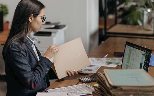 A woman in an office reviews a paper case folder beside a tall stack of aged file records and a laptop