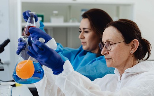 Two scientists in gowns and blue gloves examine a flask of colored liquid in a lab