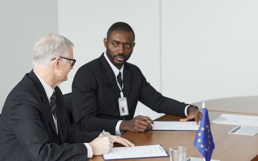 Two officials in dark suits sign documents at a conference table with a European Union flag between them