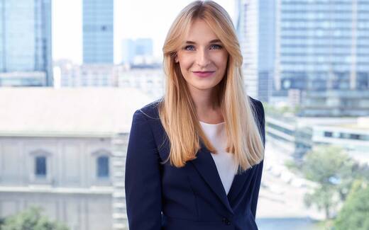 A businesswoman in a navy blazer smiles in an office with a city skyline visible through the window behind her