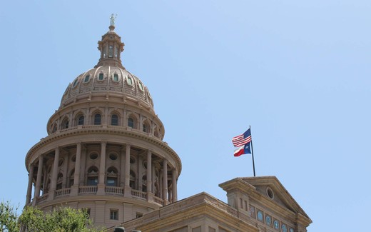 The Texas State Capitol dome against a clear blue sky, with the American and Texas flags flying from an adjacent rooftop