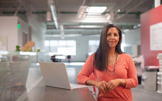 A woman stands at a desk with an open laptop in a bright, loft-style office