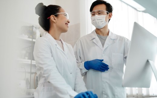 Two researchers in lab coats and safety glasses examining a specimen through a microscope