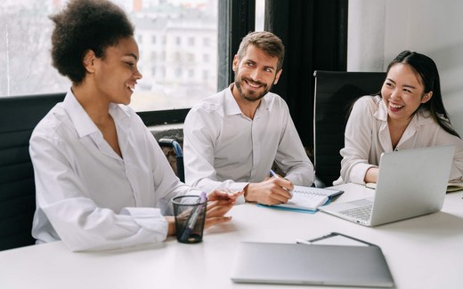 Three colleagues in white shirts share a laugh around a desk with laptops and notebooks open