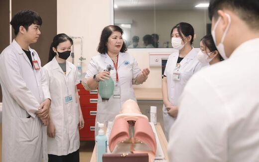 An instructor in a white coat shows a group of masked medical trainees an airway manikin in a clinical training room, a generic stand-in for clinical and medical-education settings