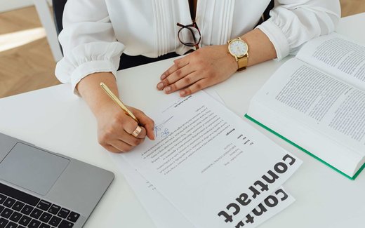 A close-up of a hand signing a contract document with a gold pen next to a laptop