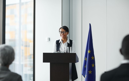 A woman in glasses smiles while speaking at a lectern beside a European Union flag, addressing seated attendees