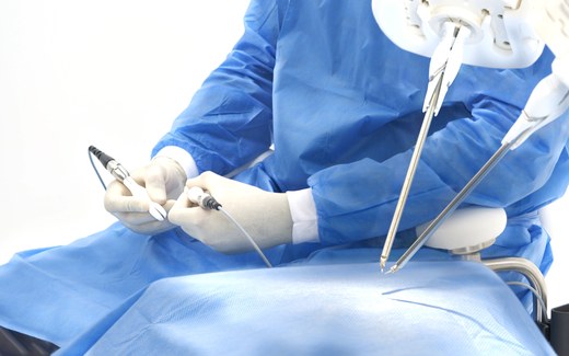 A gowned surgeon holds the Symani hand controllers as two robotic microsurgery instruments reach toward the draped field