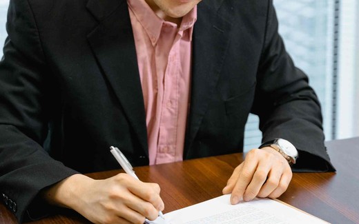 A close-up of a man in a suit signing a printed document with a pen at a wooden desk