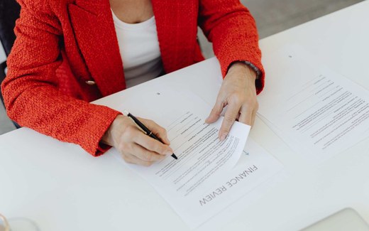 A person in a red blazer signs a paper form on a desk, pen in hand