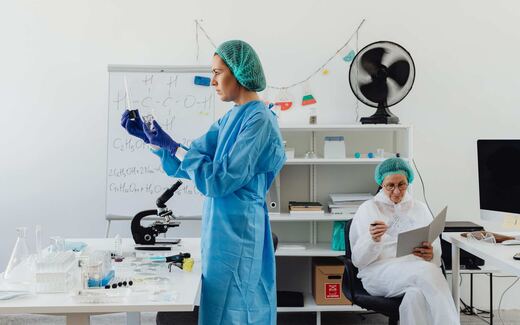 A scientist in a lab gown examines samples at a lab bench with a microscope and glassware