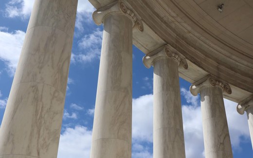 Marble columns and rotunda ceiling of a neoclassical government building, viewed from below against a blue sky