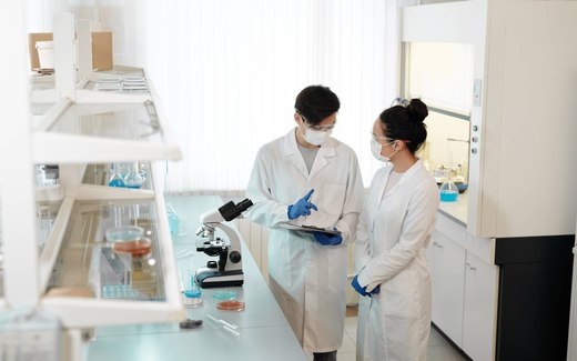 Two masked researchers in white lab coats confer over a clipboard in a laboratory, standing beside a microscope and glassware