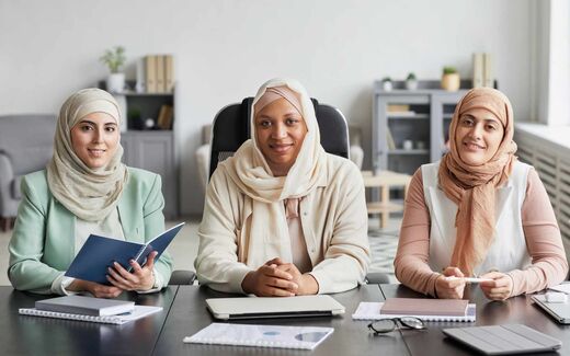 Three colleagues in business attire sit together at a conference table reviewing documents in a bright office