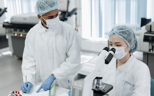 Two gowned, masked laboratory researchers in hairnets examining a sample under a microscope while taking notes