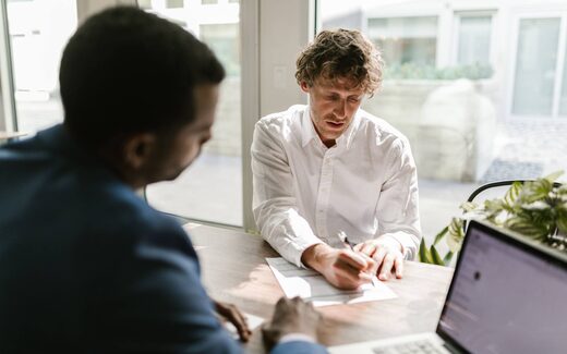 A man in a white shirt reviews and signs a document at a table near a bright window