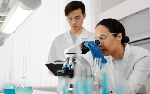 A researcher examines a specimen through a microscope while a colleague takes notes on a tablet in a bright laboratory