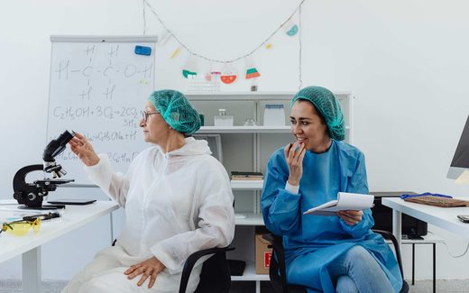 Two scientists in a laboratory, one gesturing toward a microscope while the other looks on and smiles
