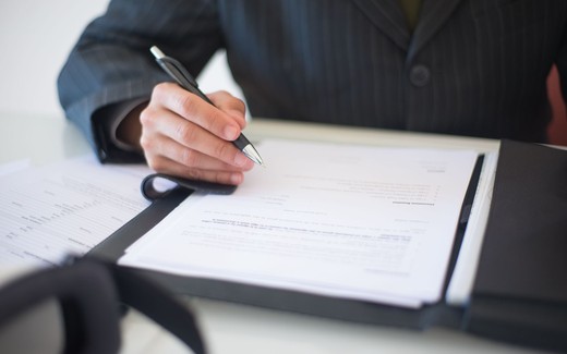 A close-up of a hand in a pinstripe suit signing a regulatory document on a clipboard