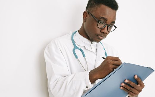 A physician in a white coat with a stethoscope writes notes on a folder against a plain studio background