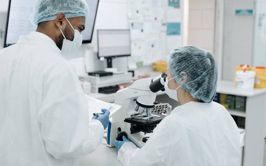 Two gowned technicians in hairnets and masks conferring over a microscope and lab notes in a cleanroom