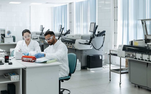 Two lab technicians in a clinical laboratory review blood sample vials and analyzer readouts