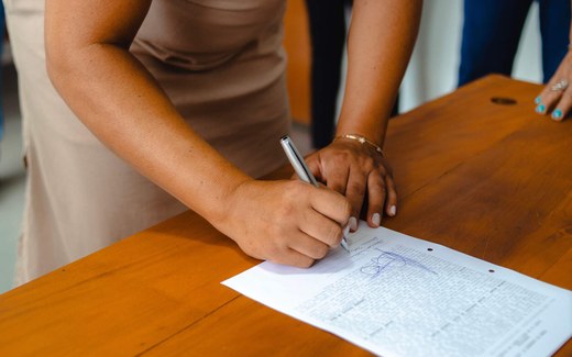 A person signs a printed document by hand with a pen while a colleague stands nearby at a wooden table