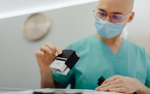 A masked clinician in scrubs examines a handheld stamping device over paperwork on an exam-room counter