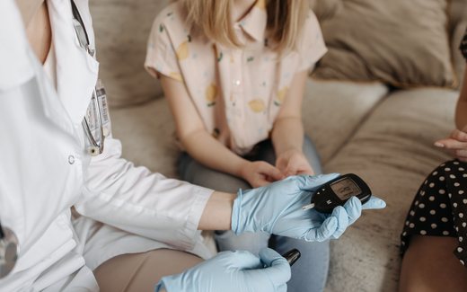 A physician in a white coat and gloves checks a patient’s blood glucose with a meter during a consultation