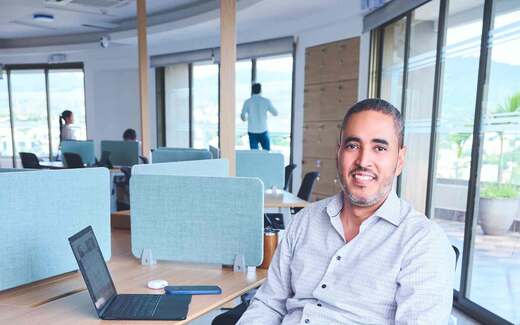 A solo executive in a checked shirt smiling at his desk in a bright, glass-walled open office