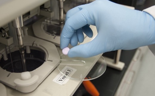 A gloved hand holding a tablet over dissolution-testing equipment in an FDA laboratory