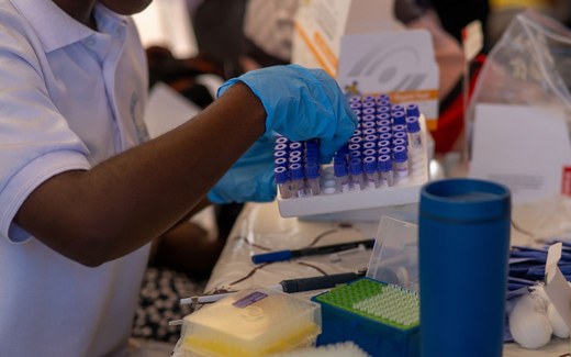 A gloved hand lifts a rack of purple-capped sample collection tubes at a testing table, with a rapid-test box in the background