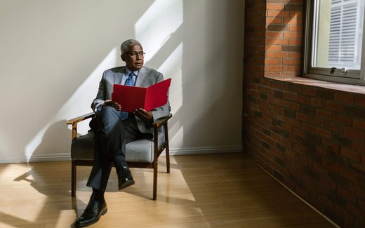 A man in a grey suit sits in a sunlit chair reading from an open red folder, near a window and a brick wall