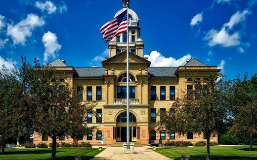 A stone government building with a domed clock tower and a large American flag flying from a pole out front, under a bright blue sky