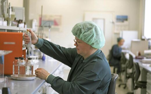 A lab technician in a hairnet and gloves pipetting samples at a bright laboratory bench
