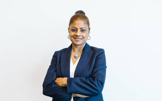A woman in a dark blazer stands with arms crossed against a plain white wall