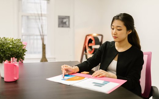 A professional in a dark blazer reviews a colorful bar and pie chart report at a desk in a bright office