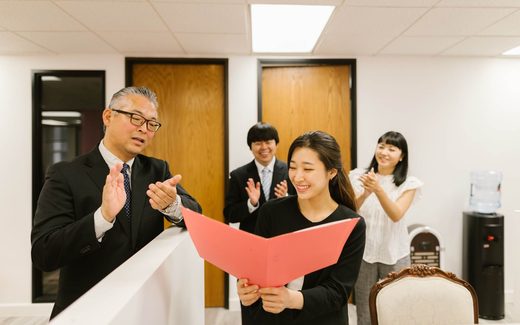 Colleagues applaud as one holds open a red folder in an office setting, marking an official milestone