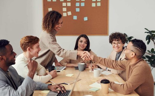 A diverse group of colleagues around a shared table, two shaking hands and smiling