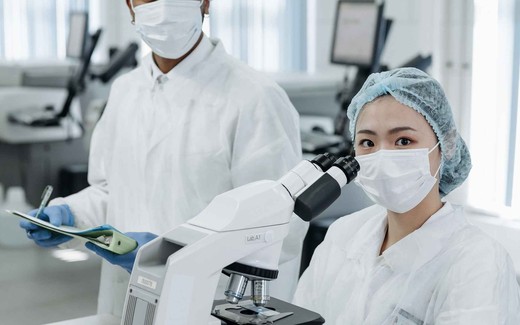 Two masked, gowned laboratory researchers in a cleanroom review a sample slide loaded on a light microscope