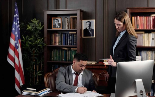 A man in a suit signs paperwork at a desk while a colleague reviews a document beside him in a wood-paneled office with an American flag