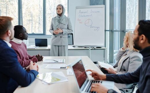 A presenter in business dress stands beside a flip chart headed Annual Income Statement while colleagues review documents at a boardroom table