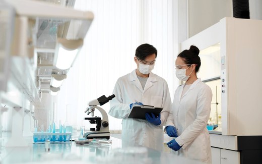 Two masked lab researchers in white coats and safety glasses review notes on a clipboard beside a microscope