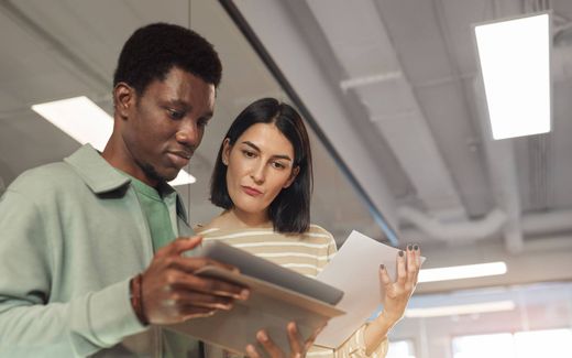 Two colleagues stand together reviewing a document and a tablet in a bright office