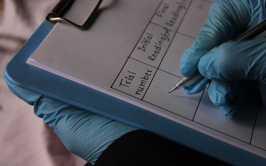 A gloved hand records readings on a clinical trial data sheet on a clipboard