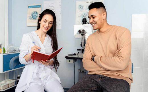 A clinician and patient smile together during an eye exam, with an eye-anatomy chart and slit lamp in the background