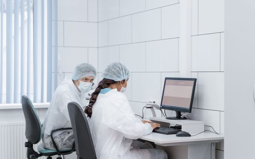 Two researchers in cleanroom coveralls, hairnets and masks review data on a computer monitor in a lab